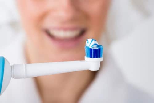 A closeup of a an electric toothbrush with toothpaste on top, woman blurry in the background