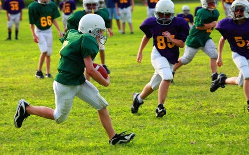 Young boys running on grass with a football, wearing helmets and jerseys