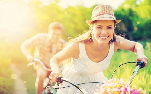 Young woman wearing a hat, smiling and riding a bike with a man following behind her