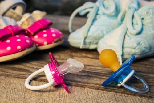 One pink pacifier and one blue pacifier with two pairs of pink and blue shoes behind them